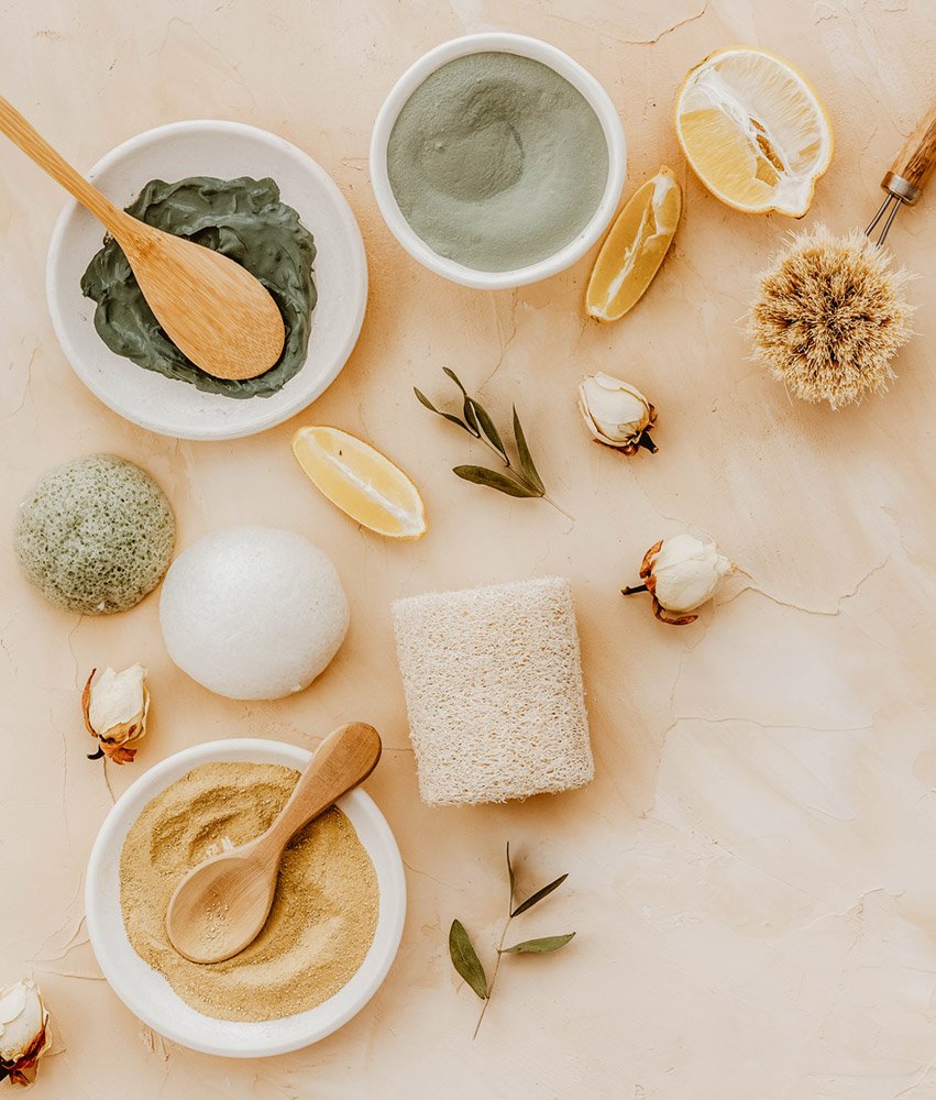 Vegane Kosmetikprodukte, darunter grüne und graue Masken, Peeling-Schwämme, Zitrusfrüchte und natürliche Inhaltsstoffe, auf einem hellen Untergrund, umgeben von Pflanzen und Rosenblüten.