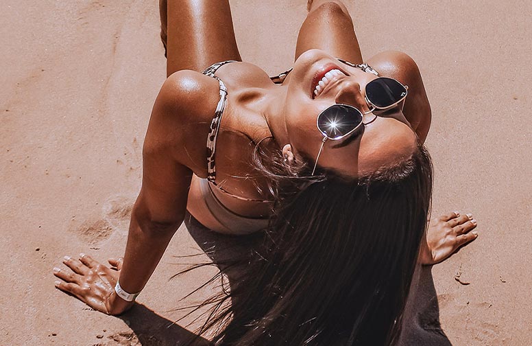 Sommer-braeune-02 Frau mit Sonnenbrille und sonnengebräunter Haut, lächelnd am Strand, symbolisiert Sommerurlaub und Hautpflege für langanhaltende Bräune.