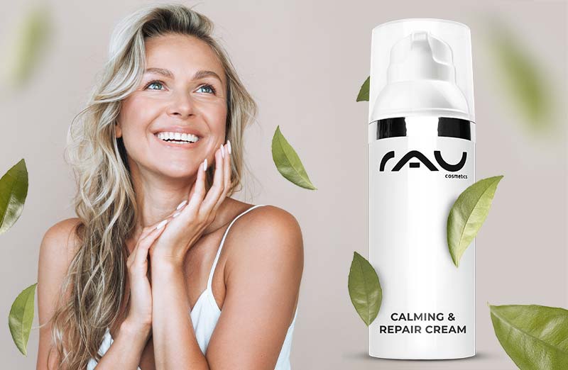 Blonde Frau trägt die RAU Cosmetics Calming & Repair Cream auf, im Hintergrund sind zarte grüne Blätter zu sehen.