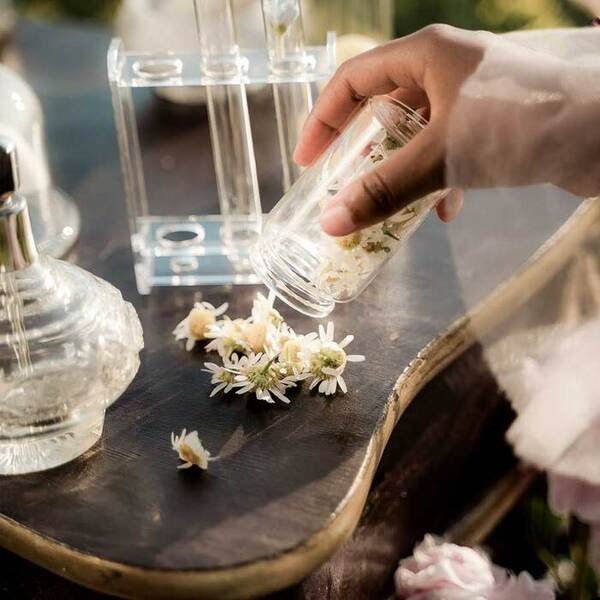 Hände einer Person mit einem Glasgefäß, frischen Blumen und Parfümflaschen für RAU Cosmetics Hautpflegeprodukte.