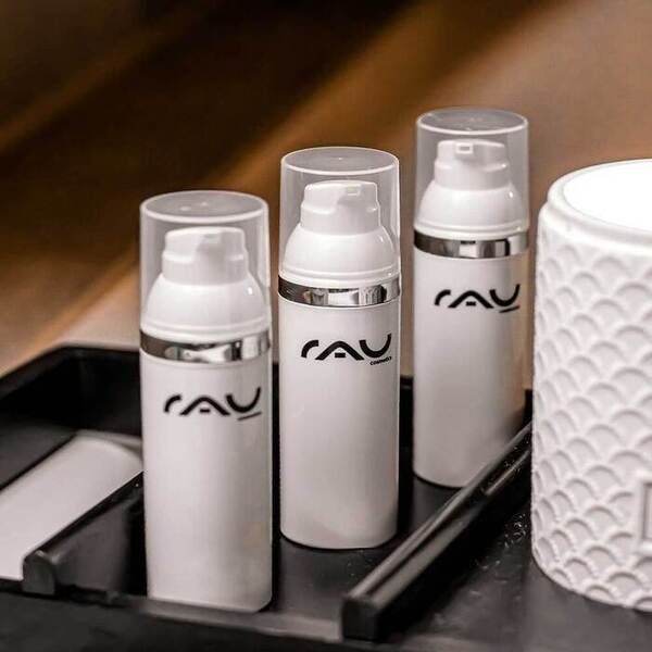 Drei weiße Hautpflegeflaschen mit RAU-Logo auf einem Tablett, neben einer dekorativen weißen Dose, präsentiert als Teil einer hochwertigen Wirkstoffkosmetiklinie.