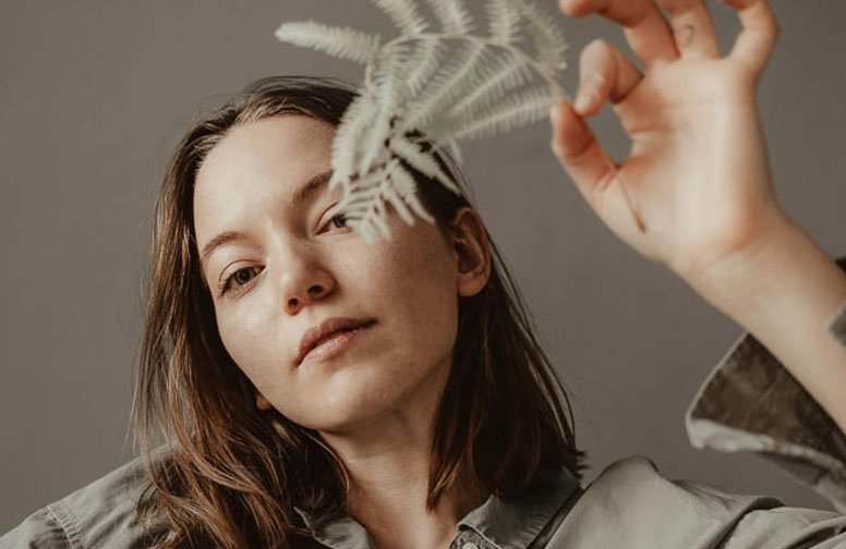 Frau mit leichtem Make-up und braunen Haaren, die ein Blatt hält, symbolisiert natürliche Hautpflege und betont die Bedeutung von Feuchtigkeit für die Haut im Winter.