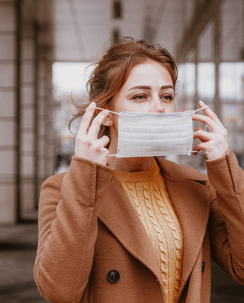 Frau in braunem Mantel und gelbem Pullover setzt eine Gesichtsmaske auf, symbolisiert die Herausforderungen von Maskenpflicht und Hautpflege während der Pandemie.