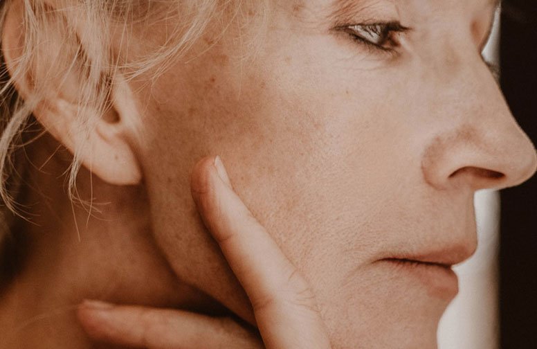 Frau mit reifer Mischhaut, die ihren Gesichtsausdruck betrachtet und sanft ihre Wange berührt, symbolisiert die Suche nach effektiven Anti-Aging-Lösungen.