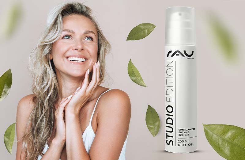 Frau mit strahlendem Lächeln und gesunder Haut, präsentiert das RAU Sunflower Enzyme Peeling in einer eleganten Verpackung, umgeben von grünen Blättern, symbolisiert sanfte Hautpflege und Anti-Aging-Effekte.