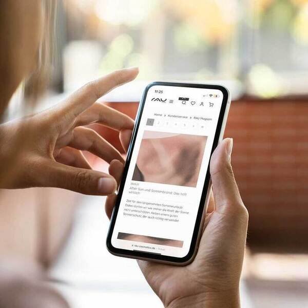 Person hält ein Smartphone, auf dem RAU Cosmetics Webseite über Sonnenlicht und Bräunung abgebildet ist.